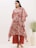 A person wearing a patterned, loose-fitting tunic with a floral design in red and beige tones. The outfit is accessorized with large, statement earrings and brown sandals. In the background, there is a white vase containing green foliage, set against a plain, light-colored wall.