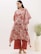A person wearing a patterned, loose-fitting tunic with a floral design in red and beige tones. The outfit is accessorized with large, statement earrings and brown sandals. In the background, there is a white vase containing green foliage, set against a plain, light-colored wall.