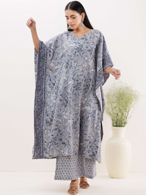 A person wearing a loose, flowing outfit with a floral pattern design, standing gracefully. The attire features a mix of soft blue and white colors, complemented by ornate earrings and sandals. In the background, there is a large white vase with green foliage, enhancing the serene atmosphere.