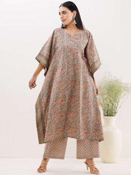 A person is wearing a flowing outfit with an intricate floral pattern in shades of orange and green on a beige base. The sleeves are wide, adding to the loose fit of the garment, paired with matching patterned pants. The individual is accessorized with large, decorative earrings and high-heeled sandals. In the background, there is a large white vase with delicate green foliage.