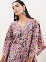 A person is wearing a floral-patterned garment with intricate designs in shades of pink, blue, and maroon. The outfit features wide sleeves and decorative trim. They have long, dark hair and are wearing large, ornate silver earrings.