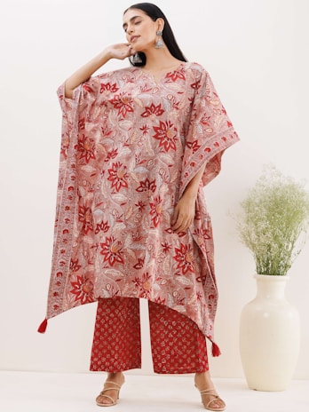 A person is wearing a flowing, floral-patterned kaftan paired with matching wide-leg trousers. The outfit features predominantly red and pink hues with intricate designs. The model is accessorized with large earrings and sandals. In the background, there is a white ceramic vase with green foliage against a neutral backdrop.
