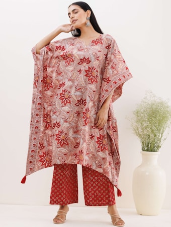 A person is wearing a flowing, floral-patterned kaftan paired with matching wide-leg trousers. The outfit features predominantly red and pink hues with intricate designs. The model is accessorized with large earrings and sandals. In the background, there is a white ceramic vase with green foliage against a neutral backdrop.