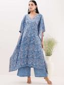 A person wearing a flowing blue kaftan with intricate floral patterns stands against a light background. The kaftan has a V-neck and is paired with matching wide-legged pants. The person is wearing sandals and large earrings, and there is a white vase with green foliage to the side.