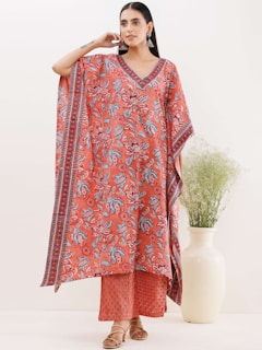 A person wearing a vibrant, floral-patterned kaftan dress stands in a relaxed pose. The dress features a mix of red, blue, and white colors with intricate floral designs. The person is accessorized with large statement earrings and sandals. A large cream-colored vase with green foliage is placed nearby, contributing to the serene ambiance.