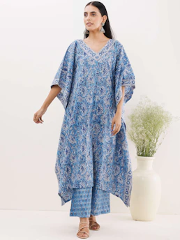 A woman is wearing a blue floral patterned kaftan with matching pants. She is accessorized with large silver earrings and nude strappy sandals. To the side, there is a tall white vase with delicate green foliage against a light background.