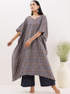 A person wears a loose-fitting, patterned dress with a geometric design in blue and reddish-brown tones. The outfit is complemented by large, dangling earrings and sandals. They stand in a relaxed pose beside a white ceramic vase containing a green plant.