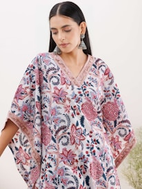 A woman wearing a floral-patterned garment with intricate designs in shades of pink, red, and blue. She is also wearing large, dangling earrings and has long, dark hair. The floral design covers the dress, which has a V-neck and wide, flowing sleeves. The background is a plain, light color.