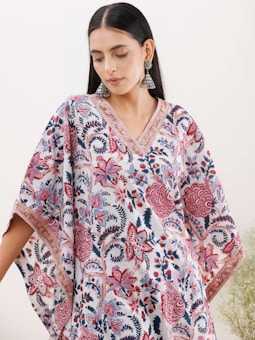 A woman wearing a floral-patterned garment with intricate designs in shades of pink, red, and blue. She is also wearing large, dangling earrings and has long, dark hair. The floral design covers the dress, which has a V-neck and wide, flowing sleeves. The background is a plain, light color.