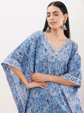 A person wearing a blue floral-patterned garment with elaborate silver earrings. The individual has long, dark hair and is posing with hands gently crossed at the waist.