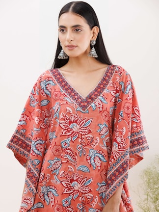 A person with long black hair wearing large jhumka earrings and a colorful garment featuring floral patterns in shades of red, blue, and white. The garment has a V-neck design with intricate patterns along the neckline and sleeves.