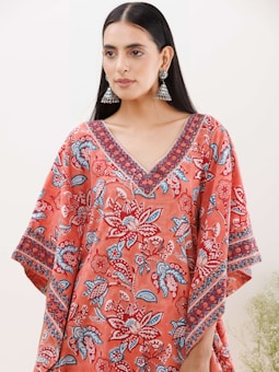 A person with long black hair wearing large jhumka earrings and a colorful garment featuring floral patterns in shades of red, blue, and white. The garment has a V-neck design with intricate patterns along the neckline and sleeves.