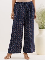 A person is wearing loose-fitting, dark blue pants adorned with a subtle floral pattern. The pants have a drawstring waist and are paired with light brown sandals. In the background, a large, cream-colored vase with green foliage adds an element of decor.