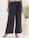 A person is wearing loose-fitting, dark blue pants adorned with a subtle floral pattern. The pants have a drawstring waist and are paired with light brown sandals. In the background, a large, cream-colored vase with green foliage adds an element of decor.