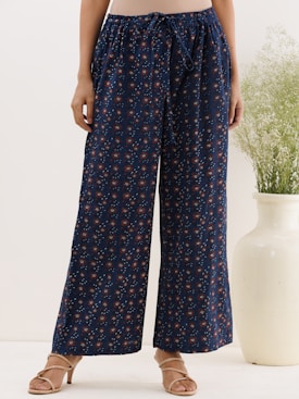 A person is wearing loose-fitting, dark blue pants adorned with a subtle floral pattern. The pants have a drawstring waist and are paired with light brown sandals. In the background, a large, cream-colored vase with green foliage adds an element of decor.