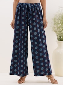 A person wearing wide-leg pants with a drawstring waist and a dark blue color featuring a light blue pattern. The individual is standing with hands relaxed at their sides, wearing beige sandals. A light, neutral background with a tall vase containing green foliage is partially visible.