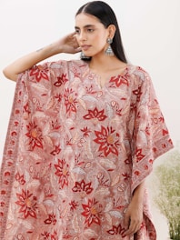 A person wearing a flowing garment with intricate floral patterns in shades of red, orange, and white. The garment features large, symmetrical flowers and leafy designs. The person is accessorized with dangling, ornate earrings and their hair is worn straight. The background is minimalistic with soft, natural light and some greenery visible at the bottom right.