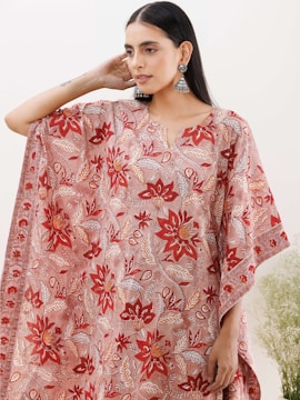 A person wearing a flowing garment with intricate floral patterns in shades of red, orange, and white. The garment features large, symmetrical flowers and leafy designs. The person is accessorized with dangling, ornate earrings and their hair is worn straight. The background is minimalistic with soft, natural light and some greenery visible at the bottom right.