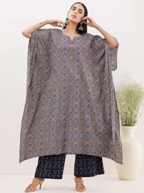 A person stands confidently wearing a loose, intricately patterned garment featuring geometric designs in shades of blue and brown. The outfit is complemented by wide-legged pants in a matching color scheme. The person accessorizes with large, dangling earrings and beige sandals. In the background, there is a tall white vase with delicate greenery, set against a plain, light-colored wall.