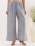 A person is wearing wide-legged pants with an intricate blue and white floral pattern. The pants have an elastic waistband with a drawstring. The outfit is complemented by beige strappy sandals. A large white vase with green foliage stands beside the person.