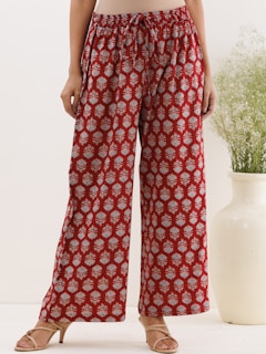 A person is wearing red wide-legged pants with an intricate white floral pattern. The pants have a drawstring waist, and the fabric appears to be lightweight. The lower body is visible, including the feet, which are wearing beige sandals with a low heel. In the background, there is a large white vase with tall, delicate green foliage.