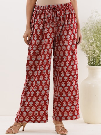 A person is wearing red wide-legged pants with an intricate white floral pattern. The pants have a drawstring waist, and the fabric appears to be lightweight. The lower body is visible, including the feet, which are wearing beige sandals with a low heel. In the background, there is a large white vase with tall, delicate green foliage.