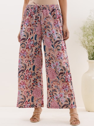 A pair of wide-leg pants with a colorful floral pattern, featuring shades of pink, red, and dark navy against a beige background. The pants have an elastic waistband with a drawstring. The outfit is paired with strappy sandals and a cream-colored top. A large white vase with green foliage is partially visible on the right.