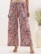 A pair of wide-leg pants with a colorful floral pattern, featuring shades of pink, red, and dark navy against a beige background. The pants have an elastic waistband with a drawstring. The outfit is paired with strappy sandals and a cream-colored top. A large white vase with green foliage is partially visible on the right.