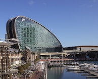 A modern, uniquely shaped glass building with a prominent 'W' letter on top, situated along a waterfront. The area features a bustling pedestrian walkway lined with shops and restaurants. Several boats are docked in the marina adjacent to the walkway. The sky is clear and blue, enhancing the vibrant atmosphere of the place.