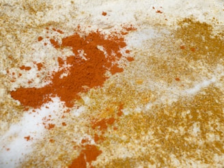 A close-up view of scattered orange and yellow powders on a white surface. The colorful powders are spread unevenly, creating a textured and dynamic contrast against the background.
