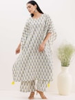 A person is wearing a flowing, white and yellow patterned kurta with matching pants. The outfit features a repeated leaf design, and the kurta has decorative yellow tassels at the bottom. The individual is standing on a neutral background with a large, cream-colored vase filled with green decorative branches beside them.