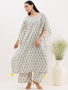 A person is wearing a flowing, white and yellow patterned kurta with matching pants. The outfit features a repeated leaf design, and the kurta has decorative yellow tassels at the bottom. The individual is standing on a neutral background with a large, cream-colored vase filled with green decorative branches beside them.