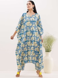 A person is wearing a loose-fitting, floral-patterned garment with shades of blue and yellow. The outfit features large, prominent flowers and flows elegantly to the floor. The person is accessorized with large, dangling earrings and is standing beside a tall white vase containing green foliage.