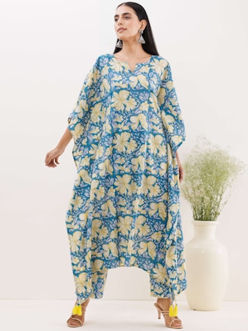A person is wearing a loose-fitting, floral-patterned garment with shades of blue and yellow. The outfit features large, prominent flowers and flows elegantly to the floor. The person is accessorized with large, dangling earrings and is standing beside a tall white vase containing green foliage.