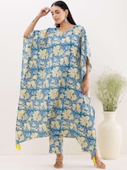A woman is wearing a flowing, floral-patterned outfit with a blue, yellow, and white design. She has long hair and is accessorized with earrings. Her sandals are light-colored, and she is standing near a large cream-colored vase with green foliage.