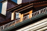 Close-up view of the architectural details of a building, focusing on a small window surrounded by wooden paneling. The roof has wooden shingles, and there is a metal railing running parallel to a green gutter.
