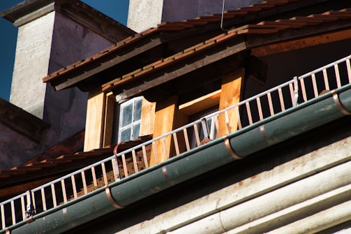 Close-up view of the architectural details of a building, focusing on a small window surrounded by wooden paneling. The roof has wooden shingles, and there is a metal railing running parallel to a green gutter.