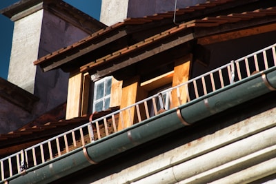 Close-up view of the architectural details of a building, focusing on a small window surrounded by wooden paneling. The roof has wooden shingles, and there is a metal railing running parallel to a green gutter.