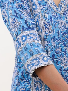 A close-up of a blue fabric featuring intricate floral and botanical patterns in shades of blue and white. The design includes swirling leaves and stylized flowers arranged in a repetitive pattern. The image shows part of a sleeve, indicating the fabric is part of a garment.