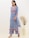 A woman wearing a blue floral-patterned kurta with intricate embroidery designs stands gracefully. She is accessorized with large silver earrings and beige heeled sandals. A large vase containing dried plants is placed beside her against a neutral-colored wall.