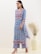 A woman wearing a blue floral-patterned kurta with intricate embroidery designs stands gracefully. She is accessorized with large silver earrings and beige heeled sandals. A large vase containing dried plants is placed beside her against a neutral-colored wall.
