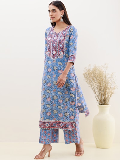 A woman wearing a blue floral-patterned kurta with intricate embroidery designs stands gracefully. She is accessorized with large silver earrings and beige heeled sandals. A large vase containing dried plants is placed beside her against a neutral-colored wall.