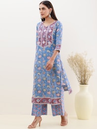 A woman wearing a blue floral-patterned kurta with intricate embroidery designs stands gracefully. She is accessorized with large silver earrings and beige heeled sandals. A large vase containing dried plants is placed beside her against a neutral-colored wall.
