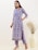 A person wearing a floral-patterned blue kurta and matching pants stands in a minimalistic setting. The kurta has a detailed collar and the person is accessorizing with large earrings and nude heels. A large beige vase with tall dried grasses is placed in the background against a neutral wall.