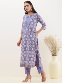 A person wearing a floral-patterned blue kurta and matching pants stands in a minimalistic setting. The kurta has a detailed collar and the person is accessorizing with large earrings and nude heels. A large beige vase with tall dried grasses is placed in the background against a neutral wall.