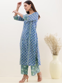 A woman wearing a blue and green patterned kurta with matching palazzo pants. She has long hair, is accessorized with earrings, and stands barefoot in a minimalistic setting. A large cream-colored vase holding dried plants is positioned beside her against a plain light background.