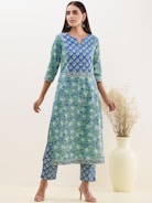 A woman wearing a blue and green floral patterned traditional outfit stands with her hands relaxed by her sides. Her attire consists of a long kurta with matching pants. She is accessorized with teal earrings and nude high heels. Beside her is a large, light-colored vase holding dried grass.