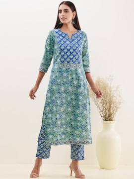 A woman wearing a blue and green floral patterned traditional outfit stands with her hands relaxed by her sides. Her attire consists of a long kurta with matching pants. She is accessorized with teal earrings and nude high heels. Beside her is a large, light-colored vase holding dried grass.