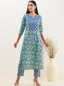 A woman wearing a blue and green floral patterned traditional outfit stands with her hands relaxed by her sides. Her attire consists of a long kurta with matching pants. She is accessorized with teal earrings and nude high heels. Beside her is a large, light-colored vase holding dried grass.