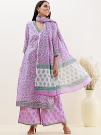 A woman is wearing a traditional Indian outfit consisting of a long, patterned kurta with matching wide-legged pants and a dupatta. The fabric is adorned with intricate floral designs in shades of purple and blue. She is accessorized with large earrings and has long dark hair. She stands next to a large white vase with dried flowers.